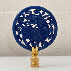 May include: A circular blue lapis lazuli lamp finial featuring an intricate openwork carving of a mythical creature. The stone disc is mounted on a polished gold-toned metal base with a threaded connector for attachment to a lamp harp.