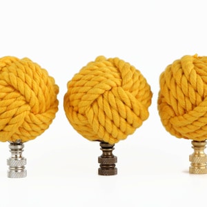 May include: Three yellow rope knot lamp finials with different metal bases. The decorative finials are made of tightly wound rope, creating a textured, spherical shape. The bases are silver, bronze, and gold.