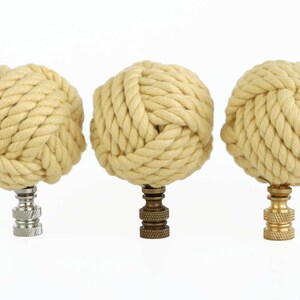 May include: Three decorative finials made of woven rope in a light beige color. Each finial is mounted on a different colored base: silver, brass, and gold.