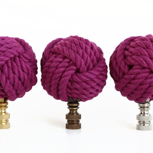 May include: Three purple rope finials with different colored bases. The finials are made of thick, braided rope and have a knot at the top. The bases are gold, bronze, and silver.