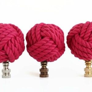 May include: Three pink rope finials with different colored metal bases. The finials are made of thick, braided rope and are tied into a knot at the top. The bases are silver, brass, and gold.