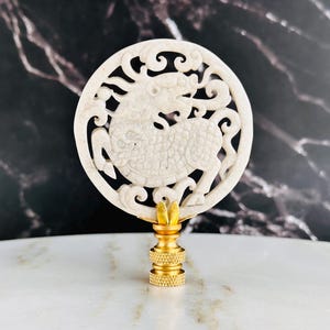 May include: A cream-colored, circular lamp finial featuring a carved design of a mythical creature within a decorative border. The finial is mounted on a gold-colored base. The background is a dark marble pattern.