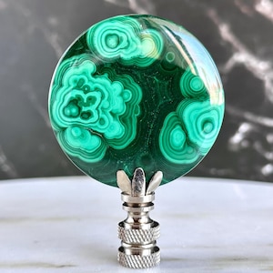 May include: A round malachite lamp finial with a polished surface. The finial features a swirling pattern of dark and light green. The base is silver-toned metal with a decorative design.