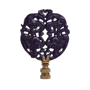 May include: A dark purple lamp finial with an intricate design featuring two stylized mythical creatures facing each other. The finial is mounted on a brass base, creating a contrast in color and texture. The design is symmetrical and detailed.