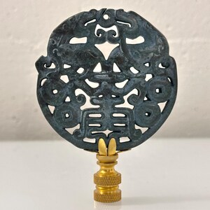 May include: A circular dark blue-grey stone finial featuring an intricate openwork carving of two symmetrical mythical creatures flanking a central character, mounted on a polished gold-toned metal base with a threaded screw attachment.