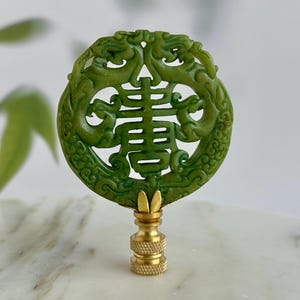 May include: A green jade lamp finial with a carved design featuring two dragons and Chinese characters. The finial has a gold-colored brass base. The jade is a rich, deep green color.