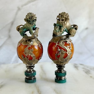 May include: Two decorative finials with orange glass globes and silver dragon and lion accents. The finials are made of metal and have a green patina.