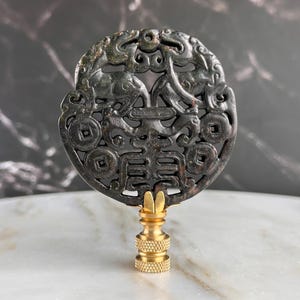 May include: A dark green, intricately carved jade pendant with a brass base. The pendant features a circular design with dragons, coins, and Chinese characters. The brass base has a textured, ridged design.