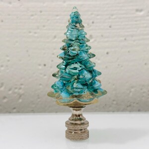 May include: A lamp finial featuring a small, conical tree sculpture made of layered, translucent turquoise and teal glass shards. The tree is mounted on a polished, metallic silver-toned base with a knurled, cylindrical neck and a threaded bottom.