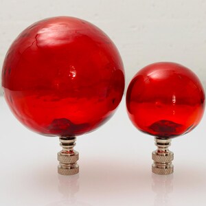 May include: Two lamp finials featuring smooth, spherical red glass orbs of different sizes. Each orb is securely mounted onto a polished silver-toned metal base with a textured, knurled finish, designed for attachment to a lamp harp.