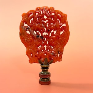 May include: A lamp finial featuring a carved, translucent orange stone ornament with intricate, symmetrical swirling patterns. The stone is mounted on a tiered, metallic bronze-colored base with a knurled texture on the bottom cylindrical section.