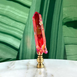 Könnte beinhalten: Ein rosa Kristall-Lampenaufsatz mit einer goldenen Metallbasis. Der Kristall ist klar mit einem rosa Farbton und hat ein facettierendes Design.