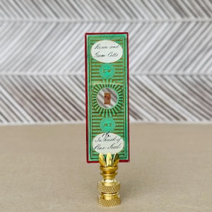 May include: A vintage lamp finial featuring a brass base and a rectangular label in green and red. The label displays the text "Resin and Gum Cells" and "In husk of Pine Seed". The finial is topped with a decorative, gold-coloured element.