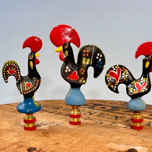 May include: Three lamp finials featuring black rooster figures with bright red combs, yellow beaks, and colorful heart and dot patterns. Each rooster stands on a light blue base attached to a brass-colored metal fitting with red rings.