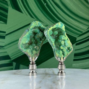 May include: Two green agate crystal lamp finials with a silver base. The crystals are a light green with a yellow hue and have a sparkly finish.