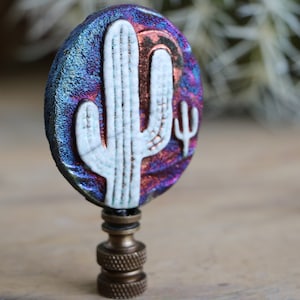 May include: A decorative lamp finial featuring a white cactus design on a dichroic glass oval. The glass has a shimmering blue, purple, and red color. The finial is attached to a brass base.