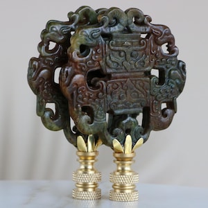 May include: A pair of ornate lamp finials made of green and brown jade with intricate carvings. The finials are mounted on gold-colored bases.