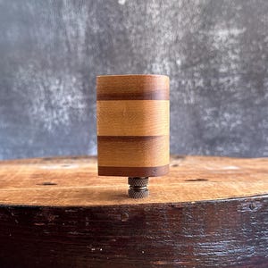 May include: A wooden guitar tuning peg with a brass base. The peg is made of multiple wood strips in different shades of brown.