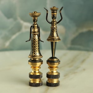 May include: Two black and gold decorative lamp finials shaped like stylized figures. The figures are wearing elaborate headdresses and clothing with gold accents.