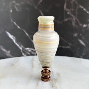 Pode incluir: Um remate de lâmpada decorativo com formato de vaso. O remate é feito de uma pedra listrada verde claro e branca, com uma base de metal cor de bronze. A parte superior do remate tem um formato alargado com uma cor verde claro.
