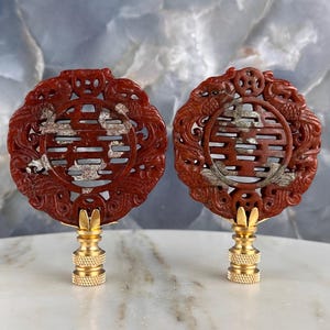 May include: Two red carved lamp finials with a circular design featuring a Chinese symbol and dragon motifs. The finials have gold-colored bases and are displayed on a white surface.