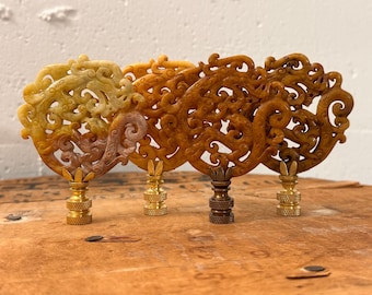 May include: Four lamp finials featuring circular, intricately carved stone ornaments in varying shades of yellow, amber, and brown. Each piece displays a swirling, openwork pattern and is mounted on a metallic base with a threaded connector.