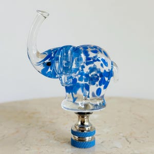 May include: A clear glass elephant bottle stopper with blue and white speckled accents. The elephant has a raised trunk and sits atop a silver and blue metal base. A decorative item.