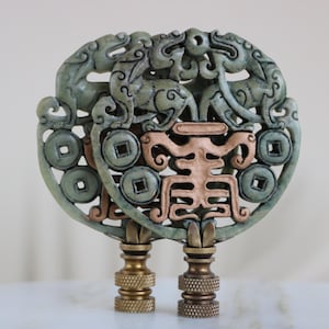 May include: Two decorative lamp finials with an Asian-inspired design. The finials feature a jade-green, openwork design with dragons, coins, and a central copper-colored symbol. The bases are brass-toned with textured detailing.