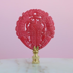 May include: A red, carved, circular lamp finial with intricate designs. The finial is mounted on a gold-coloured base.