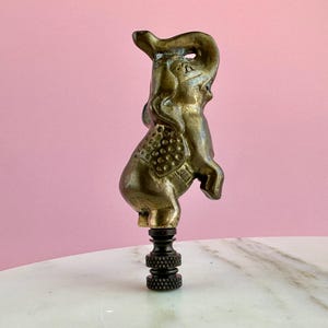 May include: A brass elephant finial with a textured surface and a raised trunk. The elephant is standing on a black metal base.