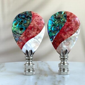 May include: Two decorative lamp finials with a teardrop shape. The finials are made of a combination of red, white, and iridescent green materials. The finials are mounted on silver bases.