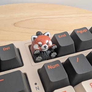 May include: A custom red panda artisan keycap sits on a computer keyboard. The keycap is a detailed, miniature sculpture of a red panda with a black and white face and a fluffy black body. The keyboard has black keys with white lettering.