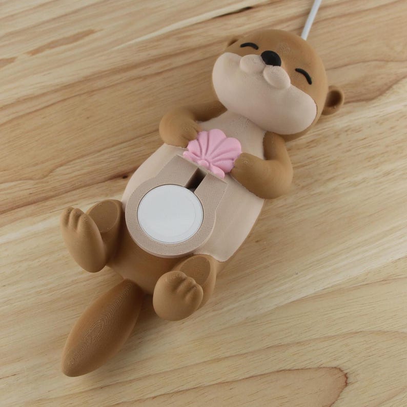 Pu&ograve; includere: Una statuetta di lontra marrone e beige con una conchiglia rosa nelle zampe. La lontra tiene un oggetto rotondo bianco nel suo ventre. La statuetta &egrave; su una superficie di legno.