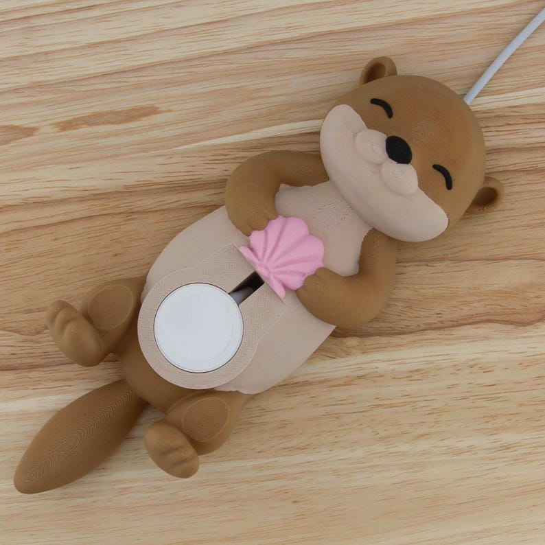 Pu&ograve; includere: Un mouse a forma di lontra di colore marrone e beige con una conchiglia rosa nelle sue mani. Il mouse &egrave; sdraiato sulla schiena con la coda arrotolata. Il mouse &egrave; collegato a un cavo bianco.