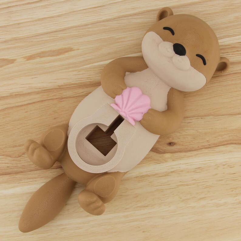 Pu&ograve; includere: Una statuetta di lontra marrone e beige con una conchiglia rosa nelle mani. La lontra ha un buco nella pancia per riporre piccoli oggetti.