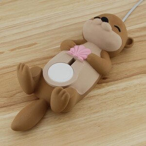 Pu&ograve; includere: Un supporto per telefono a forma di lontra marrone e beige con un dettaglio a conchiglia rosa. Il supporto ha un cerchio bianco al centro per tenere un telefono.