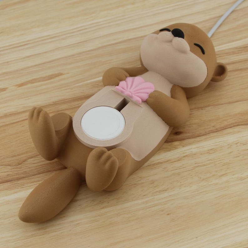 Pu&ograve; includere: Un supporto per telefono a forma di lontra marrone e beige con un dettaglio a conchiglia rosa. Il supporto ha un cerchio bianco al centro per tenere un telefono.