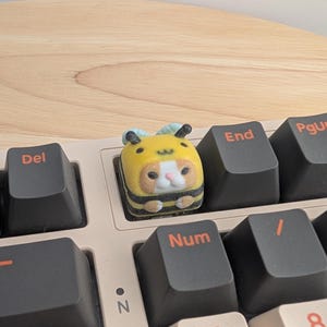 May include: A custom keycap featuring a yellow and black bee design, with a cute animal face. The keycap is installed on a mechanical keyboard with black keys and a light pink base.