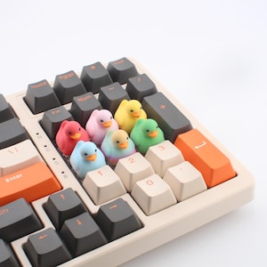 May include: A mechanical keyboard with a cream-coloured frame and dark grey and orange keycaps. Colourful rubber duck figurines are placed on the keys. The keyboard has keys labelled "Enter", "Fn", "Num", and "Pgup".