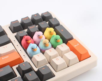 May include: A mechanical keyboard featuring a mix of dark gray, cream, and orange keycaps. Nestled between the keys are six small, colorful duck-shaped figures in red, pink, yellow, green, blue, and a multi-colored pastel gradient finish.