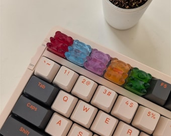 May include: A mechanical keyboard with colorful gummy bear keycaps. The keycaps are red, blue, purple, orange, and green. The keyboard has white and gray keys, with a small succulent plant in a white pot in the background.