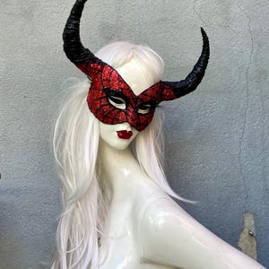 May include: A red and black glitter mask with curved horns, displayed on a mannequin with white hair. The mask has a spiderweb design and is paired with red lipstick. The horns are black and curved.