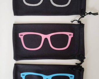 May include: Three rectangular black drawstring pouches arranged vertically. Each pouch features a graphic of thick-rimmed glasses on the front. The top pouch has silver glitter glasses, the middle has pink glasses, and the bottom has light blue glasses.