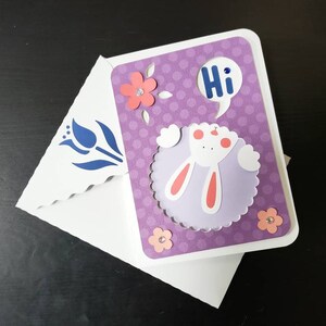 May include: A handmade greeting card with a purple polka dot background and a white bunny design. The card says "Hi" in a speech bubble. Pink flowers and rhinestones decorate the card, which is inside a white envelope with a blue floral design.