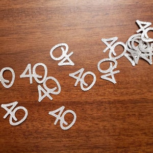 May include: Silver glitter confetti with the number 40 printed on each piece. The confetti is scattered on a brown wooden surface.