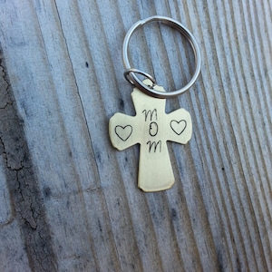 May include: A gold-toned cross keychain with the word "MOM" engraved on it. Two small hearts are engraved on either side of the word. The keychain is attached to a silver key ring.
