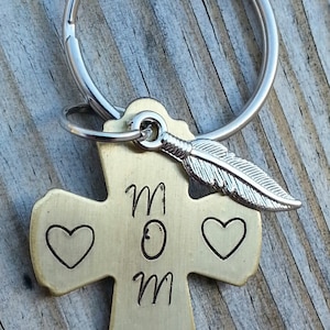 May include: A gold-toned cross keychain with a silver feather charm and the word "MOM" engraved on it. The keychain has two heart shapes on either side of the word "MOM".