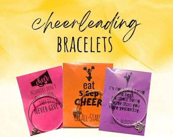 Peut inclure: Trois bracelets de cheerleading avec des citations inspirantes sur des cartes roses, orange et violettes. Les bracelets sont argentés et ont un charme de pom-pom girl.
