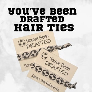 May include: Two white elastic hair ties with black soccer balls printed on them. Each hair tie has a white card attached with the text "You've Been Drafted" printed in black.