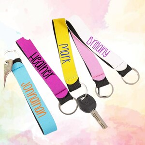 May include: Four fabric wristlet keychains with metal rings and attached keys. The wristlets are light blue with the name Jonathan, magenta with Heather, yellow with Mark, and white with Brittany printed in various colored fonts on the fabric.